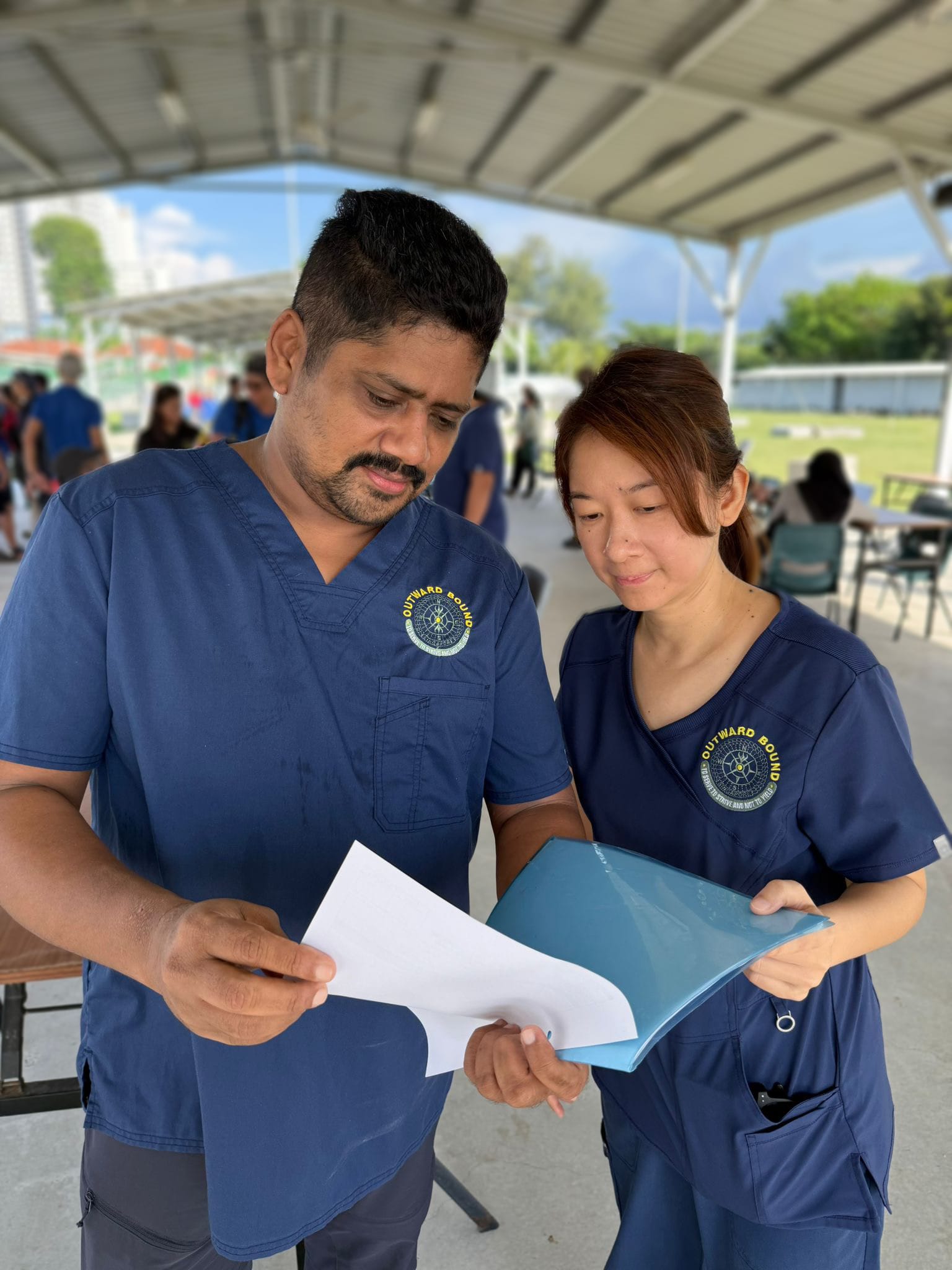 Two people in navy blue uniforms examine documents together under an outdoor shelter. 